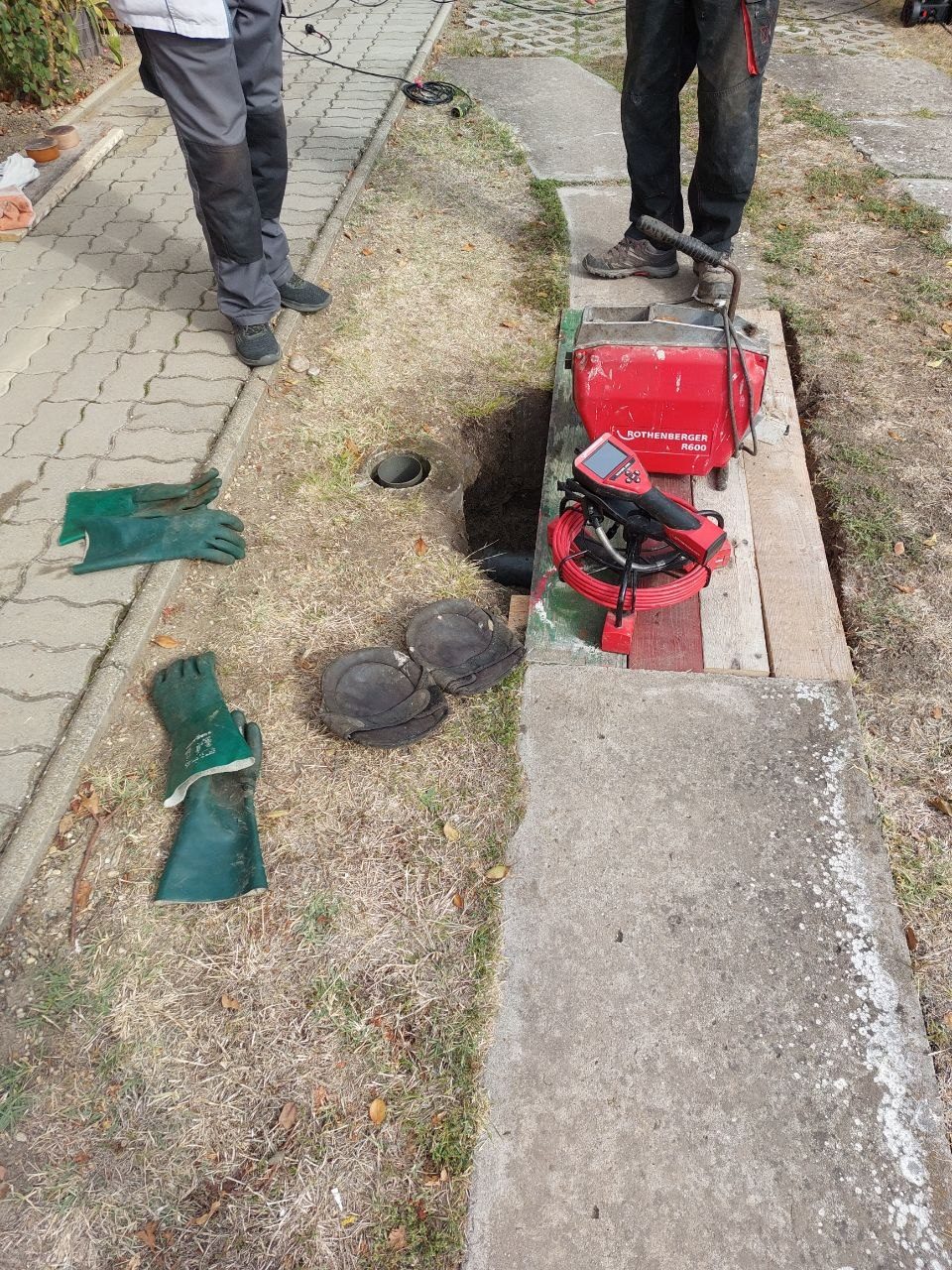 Gépi spirálos készülék deszkán az udvar füvén a földbe épített nyílás mellett, mellette védőkesztyű és két fedél