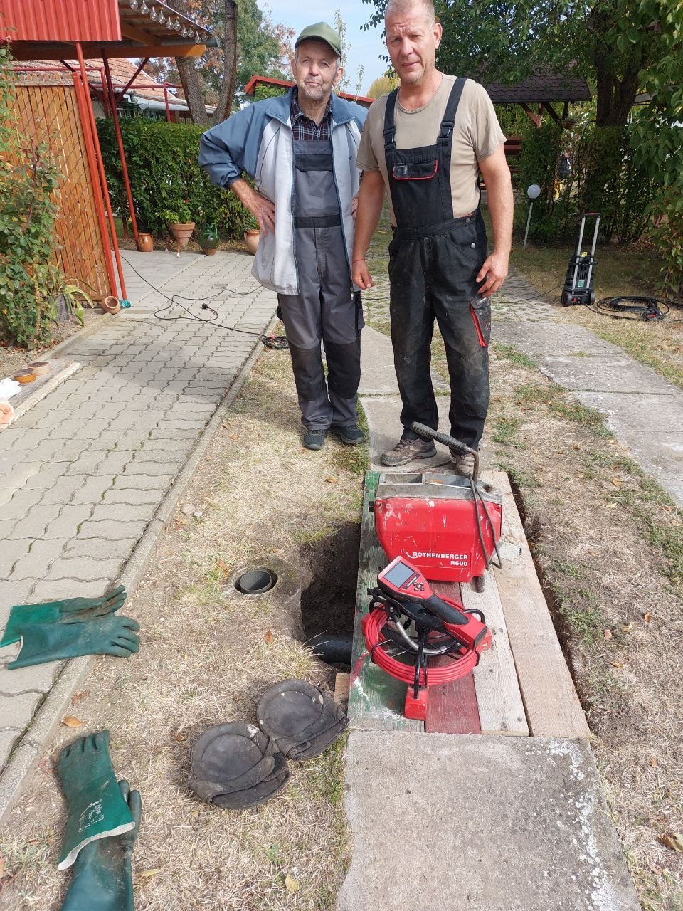 Két szerelő az udvaron a gépi spirálos készülék mögött, mosolyogva a kamerába